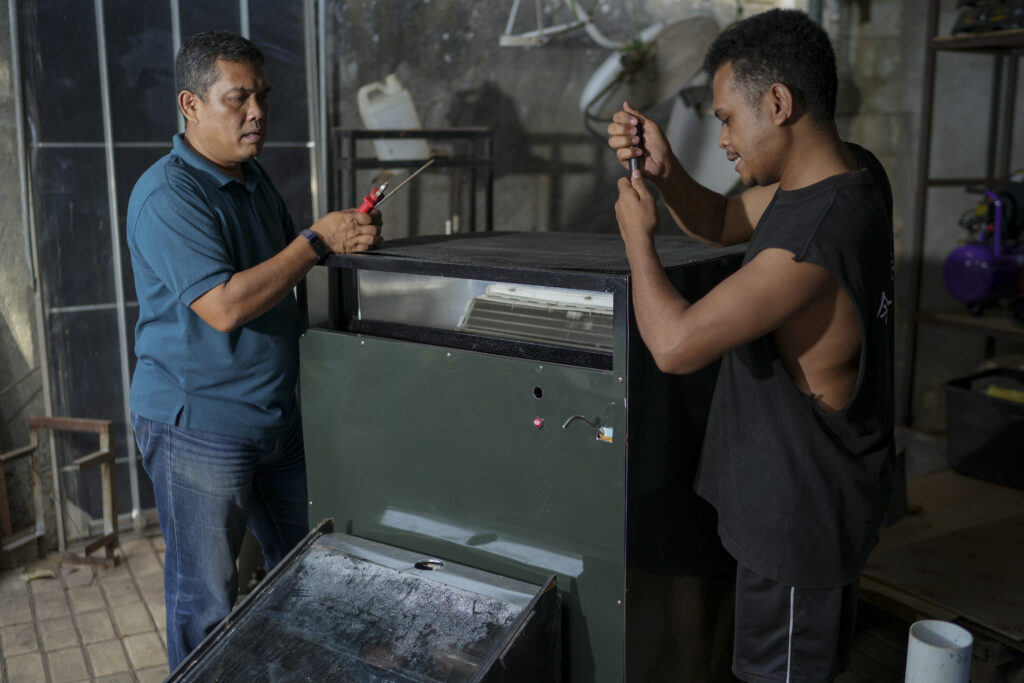 Two men work together indoors to assemble or repair a large machine, using hand tools while standing on opposite sides of the equipment in a workshop setting.