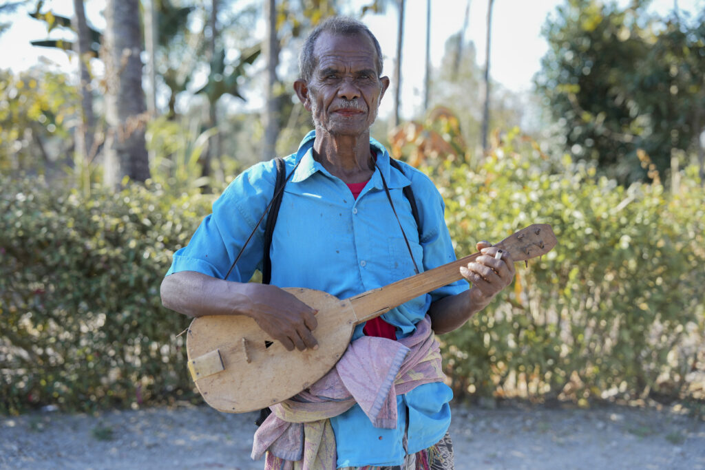 An older man stands outdoors holding and playing a small wooden string instrument, wearing a blue shirt with a cloth tied around his waist, with trees and greenery in the background.