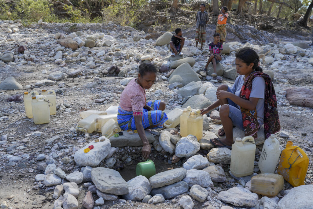 Two women collect water from a shallow pit in a dry, rocky riverbed, filling plastic jerrycans while several children sit and watch in the background.