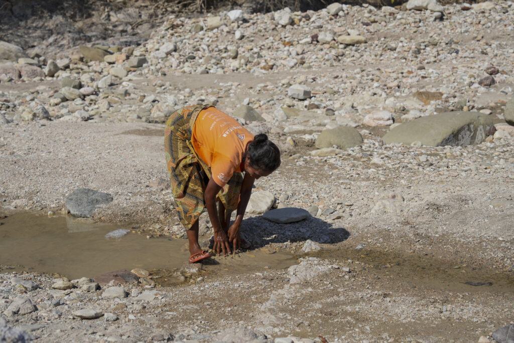A woman bends down to collect water from a small stream in a dry, rocky riverbed, using her hands amid scattered stones and shallow pools.