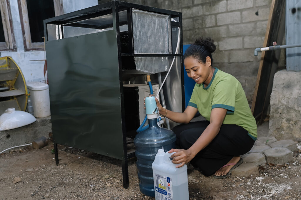 A woman crouches beside a water filtration machine, smiling as she pumps clean water from a large container into a smaller jerrycan in a simple outdoor workspace.