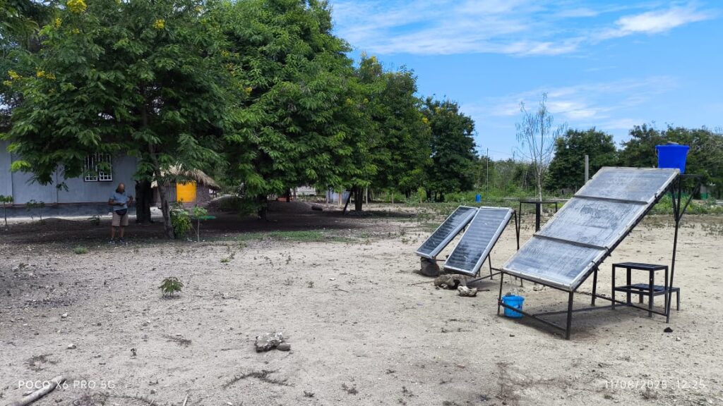 Solar panels are set up on metal frames in an open, rural yard with trees and simple buildings in the background, under a clear blue sky.