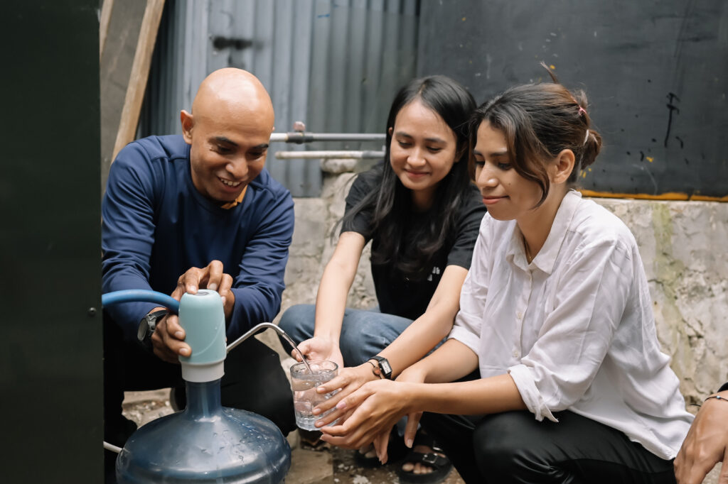 Three people crouch together while filling a glass of water from a manual pump attached to a large water container, smiling as they share the activity in a modest outdoor setting.