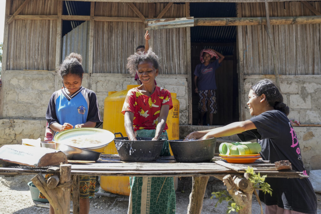Several women and a child wash dishes outdoors at a wooden table using large bowls of water, with a yellow water tank and a simple rural house in the background.