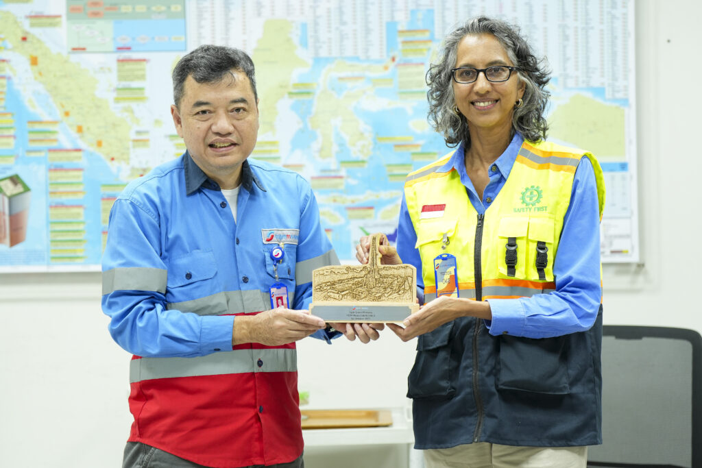 Two people (Supreme Energy President and CEO Nisriyanto and Australian Deputy Ambassador Gita Kamath) in work uniforms stand indoors in front of a wall map, smiling while holding a wooden plaque together, with one wearing a high-visibility safety vest and the other a blue industrial shirt.