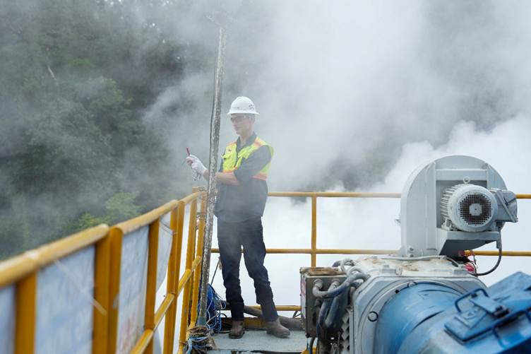 A person (David Barton) wearing a hard hat and high-visibility vest stands on an industrial platform, holding a mobile phone to photographs the steam that spins a turbine to generate electricity.